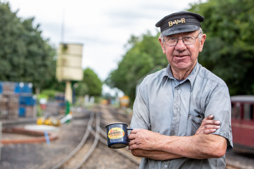 Commercial Headshot Photography of an elderly Railway worker standing on the platform wearing a Driver's cap. His clothes are dirty with smoke and oil and his arms are folded.