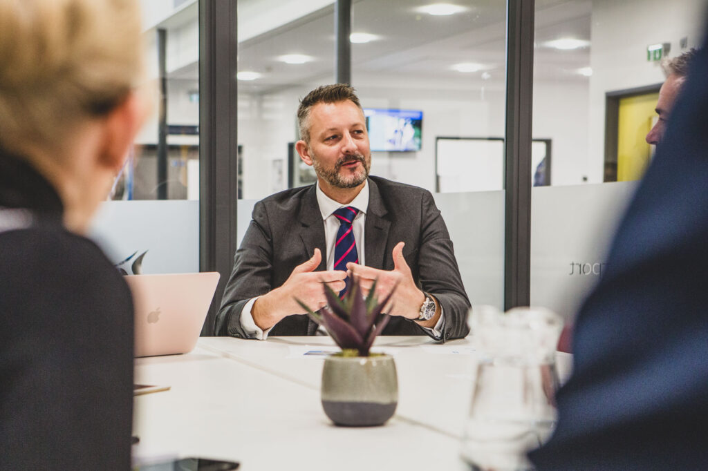 A business man in a formal suit sits at a desk in a modern meeting room addressing staff - Bespoke Stock Photography Norwich - Blanc Creative