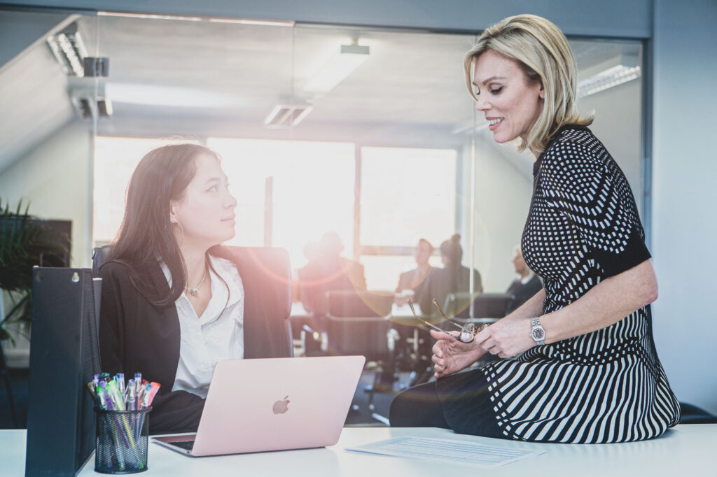 Two female office workers sit talking at a desk with sun streaming in through the window behind them. - Blanc Creative Photography Norwich