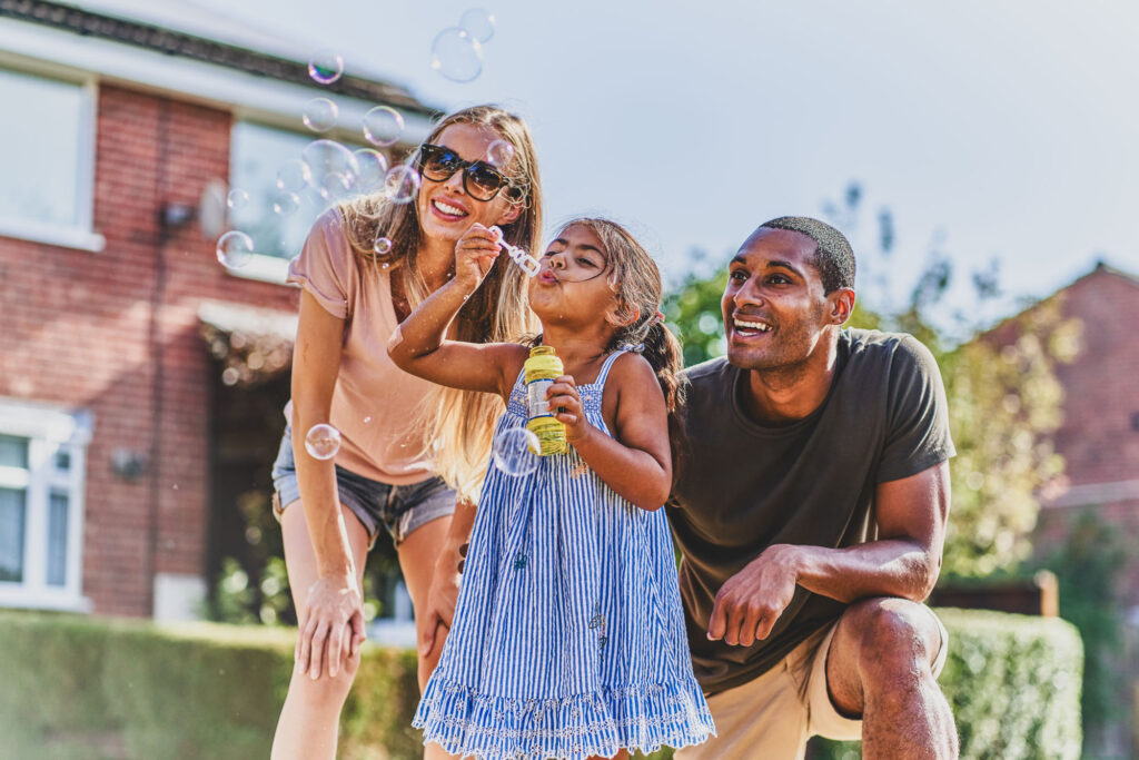 A family outside their house on a sunny day. A small girl blowing bubbles while her parents watch and smile - Bespoke Stock Photography