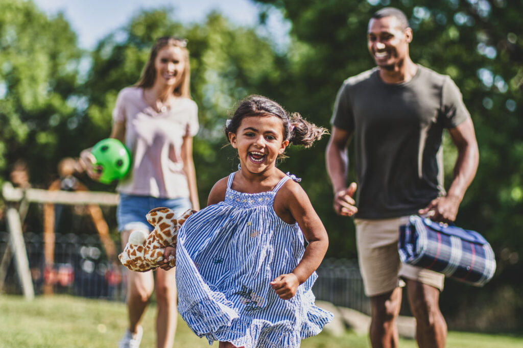 A small happy girl in a blue dress runs in a green park with a happy smile. Her parents are in the background carrying a picnic - Blanc Creative Bespoke Stock Photography