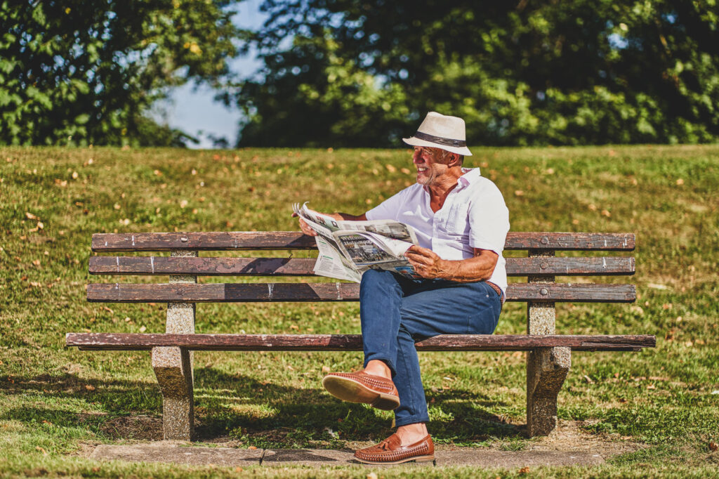 A man dressed in casual summer clothes sits soon a park bench in summer wearing a straw hat and reading a newspaper. Bespoke Stock Photography