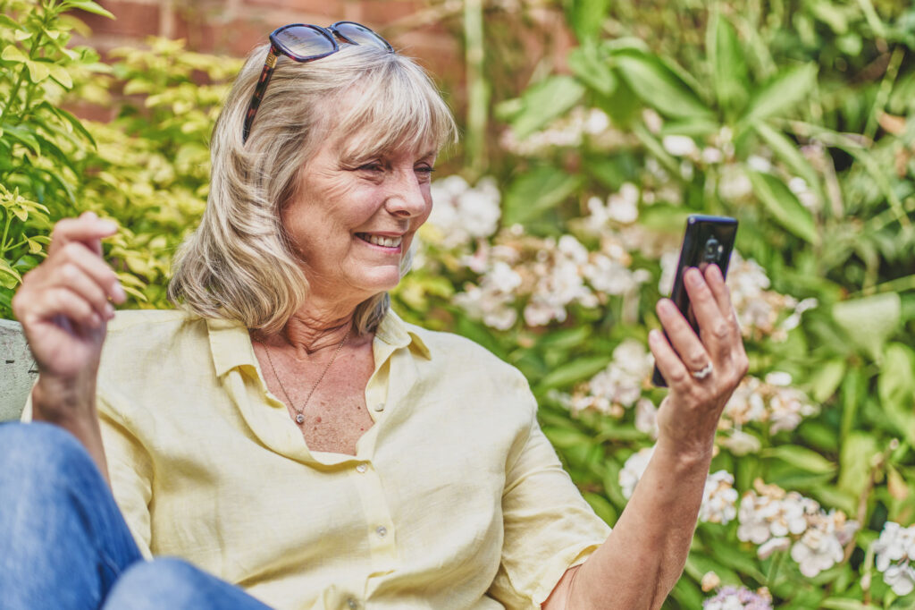 A middle aged woman sits on a bench on a sunny day and smiles as she looks at the screen of a mobile telephone - Bespoke Stock Photography Norwich