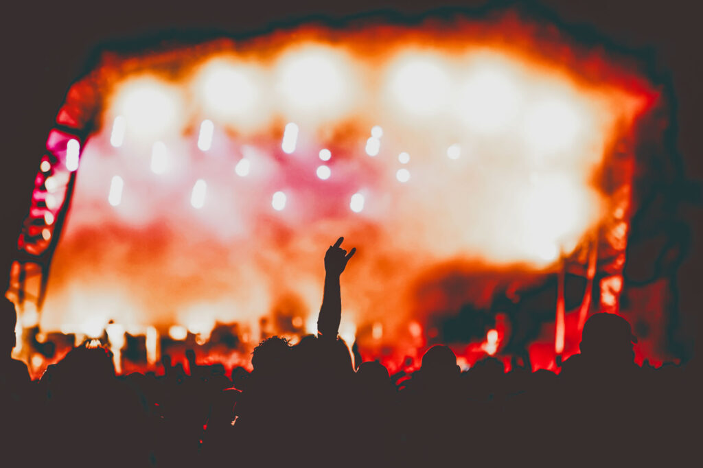 A silhouette of a concert audience against a bright orange stage. One person has their hand in the air making a love you gesture with their hand - Lee Blanchflower Bespoke Stock Photography