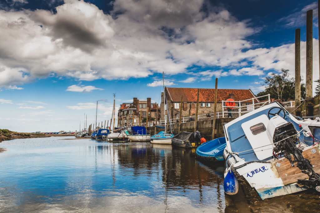 A landscape scene of a summer day at Blakeney by the Sea in Norfolk showing the sea, boats and waterfront - Photography by Blanc Creative