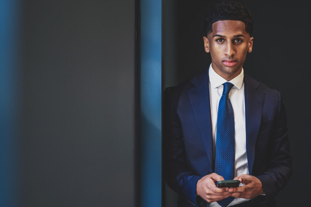 A smart young professional male wearing a well fitting business suit leans casually against a wall in a modern office