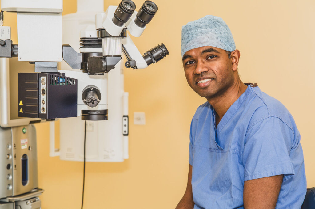 Commercial Headshot Photography Norwich showing an eye surgeon wearing scrubs sat on a stool next to a medical device - Commercial Headshot Photography Norwich