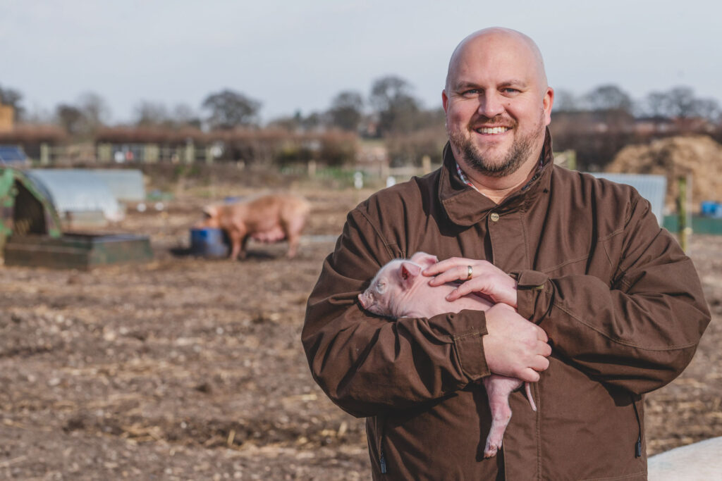 Natural Commercial Headshot Photography of Ryan Lincoln, Agricultural Partner at Lovewell Blake Chartered Accountants, standing on a farm and holding a piglet in his arms - Blanc Creative Norfolk