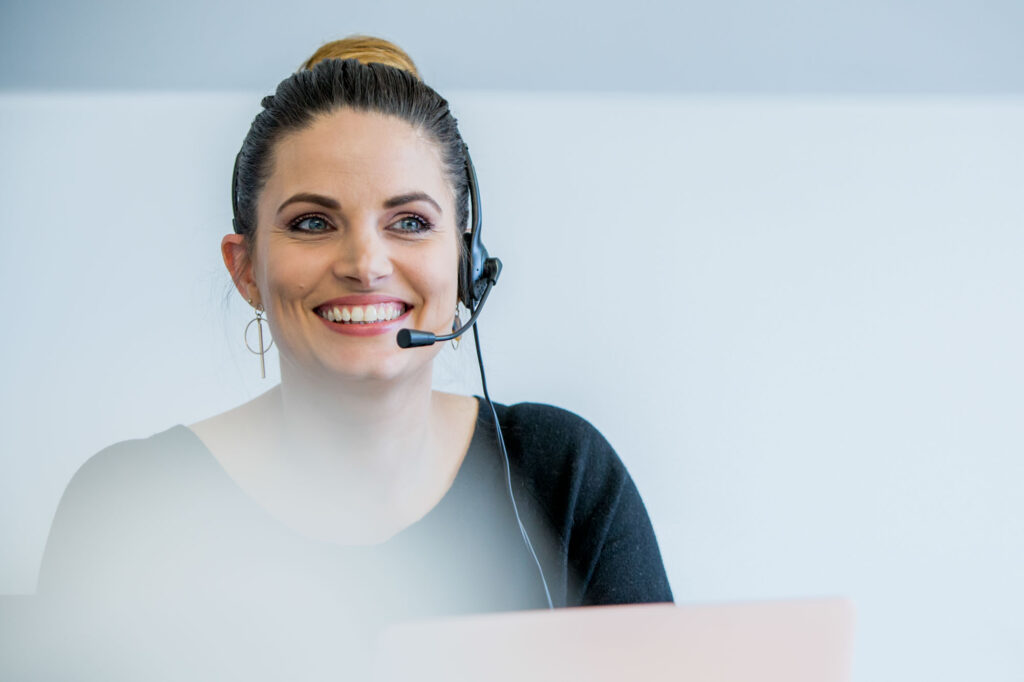 Natural Commercial Headshot Photography of a young female receptionist sits at a desk wearing a headset and smiling as she answers a telephone call - Blanc Creative Photography