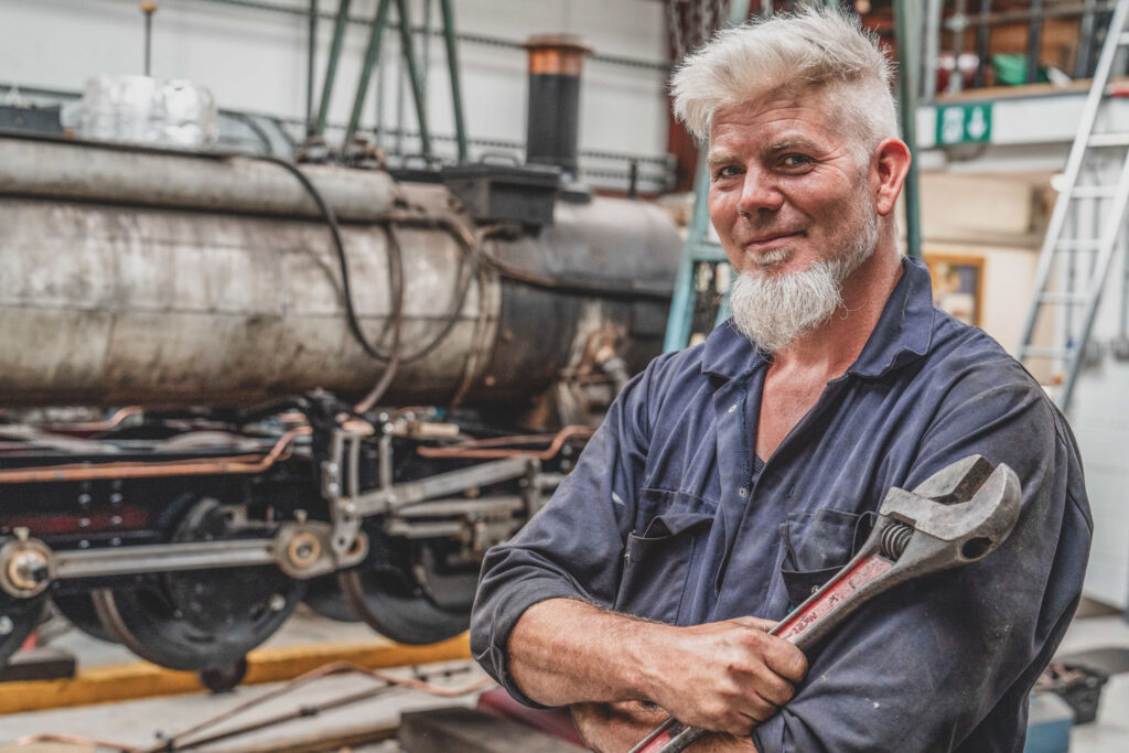 Relaxing Commercial Headshot Photography of an engineer with a grey beard and white hair standing in a workshop wearing overalls and holding a large spanner