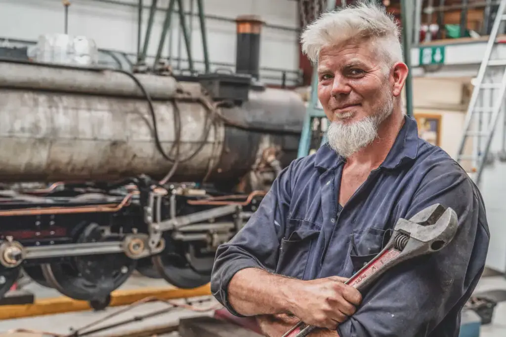 Relaxing Commercial Headshot Photography of an engineer with a grey beard and white hair standing in a workshop wearing overalls and holding a large spanner