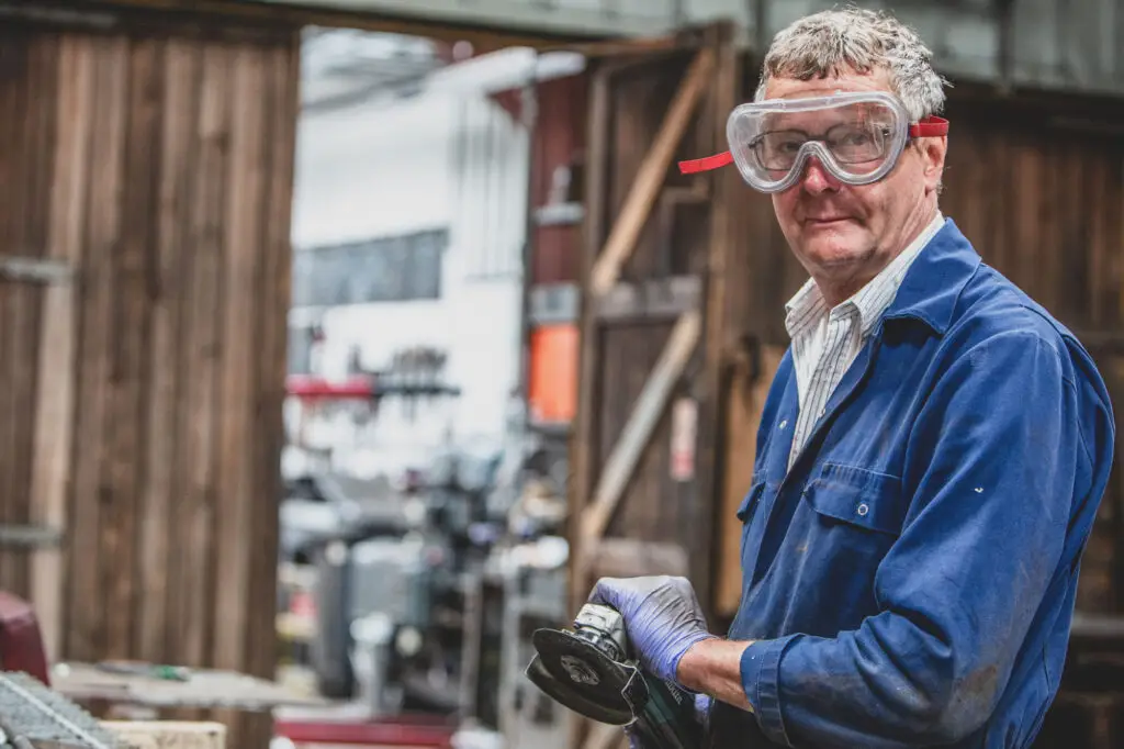 An engineers stands in a workshop at Bure Valley Railway in Norfolk holding an angle grinder and wearing overalls and safety glasses in a relaxed Commercial Headshot Photography portrait