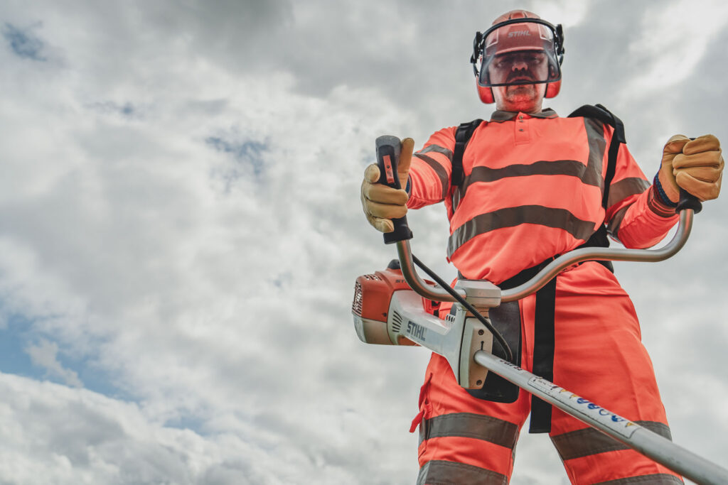 A Norse Group grounds maintenance worker holding an industrial trimmer and wearing a safety helmet, gloves and hi visibility clothing - Commercial Headshot Photography Norwich