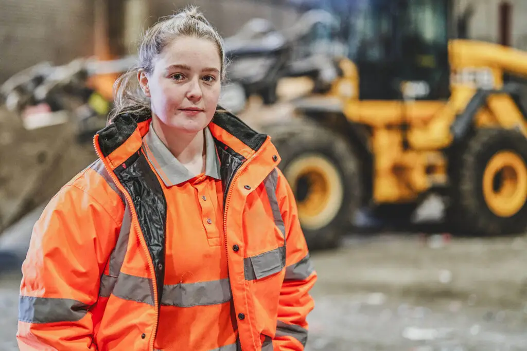 Natural Commercial Headshot Photograph of a female plant operator working in a recycling centre. Sher is wearing hi visibility clothing and standing in front of a large digger - operator