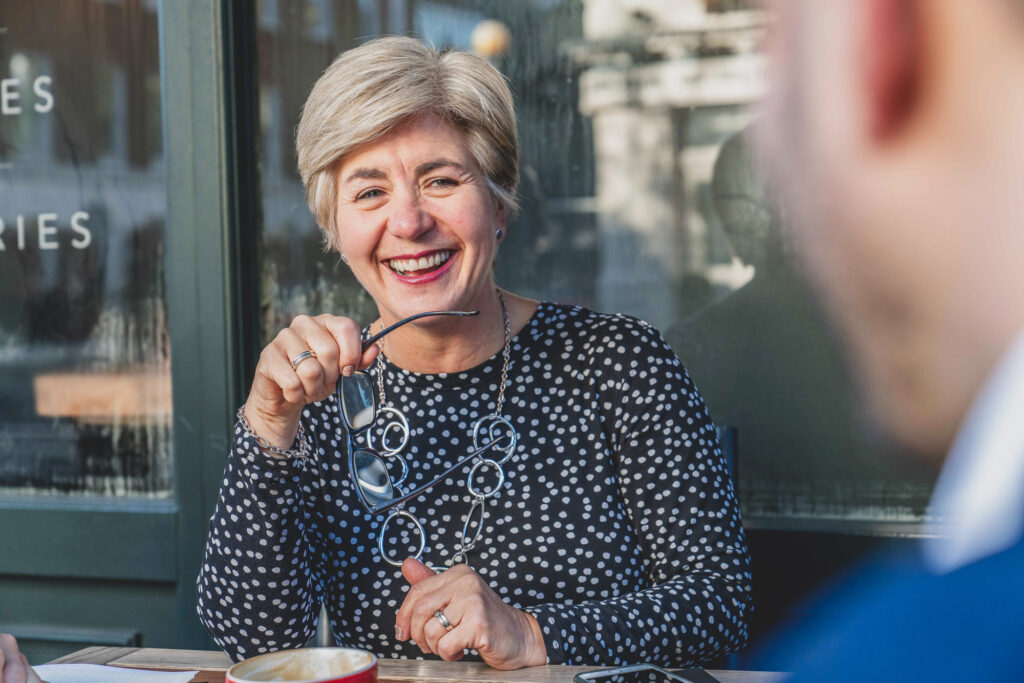 Professional Business Portraits London. A mature professional woman smiles as she looks across at a colleague during a meeting outside a coffee shop