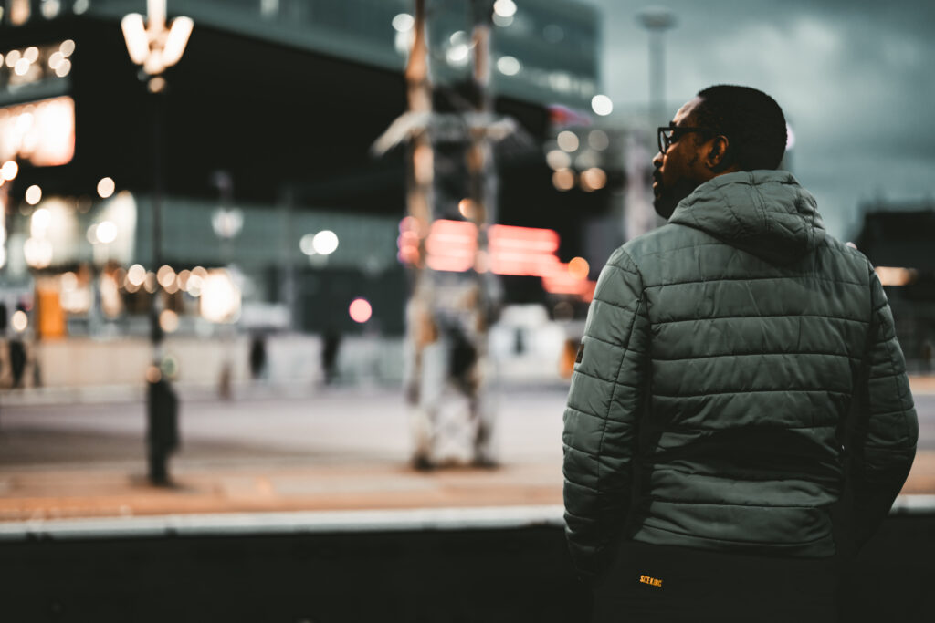 A male stands on the platform at night at Stratford station in London, waiting for a train and looking over the railway lines towards Westfield Shopping centre