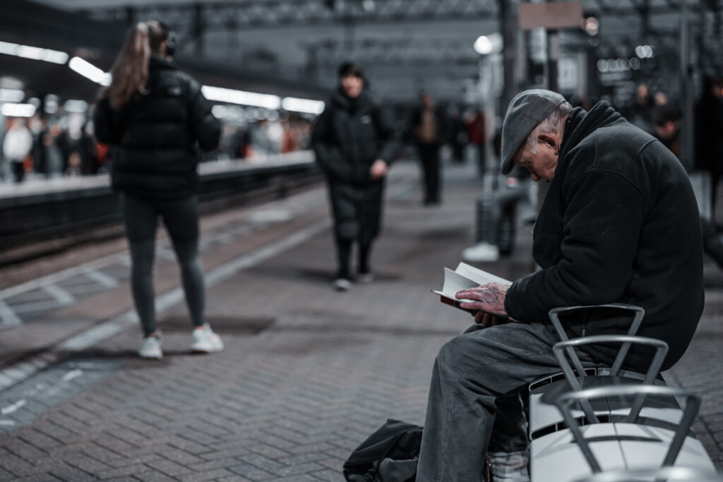 An old man sits on a bench on the platform at Stratford Station in London during rush hour and reads a book - Blanc Creative UK Photography Law